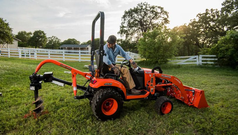 Land Pride PD10 Series Post Hole Digger mounted on a Kubota tractor near some trees and a pasture.