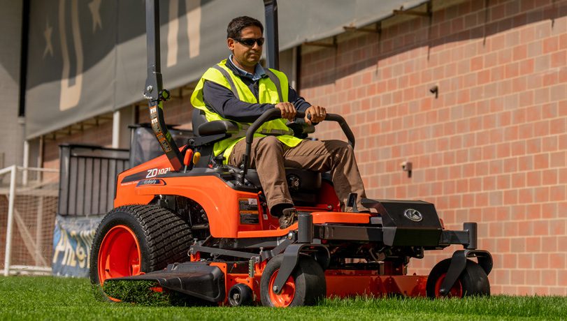 Landscaping professional driving a Kubota ZD1021-60 Mower around a large building.