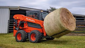 Person driving a Kubota SSV75 Skid Steer Loader moving a large bale of hay on a farm.