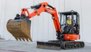 Person using a Kubota KX033 Excavator in front of a gray warehouse.