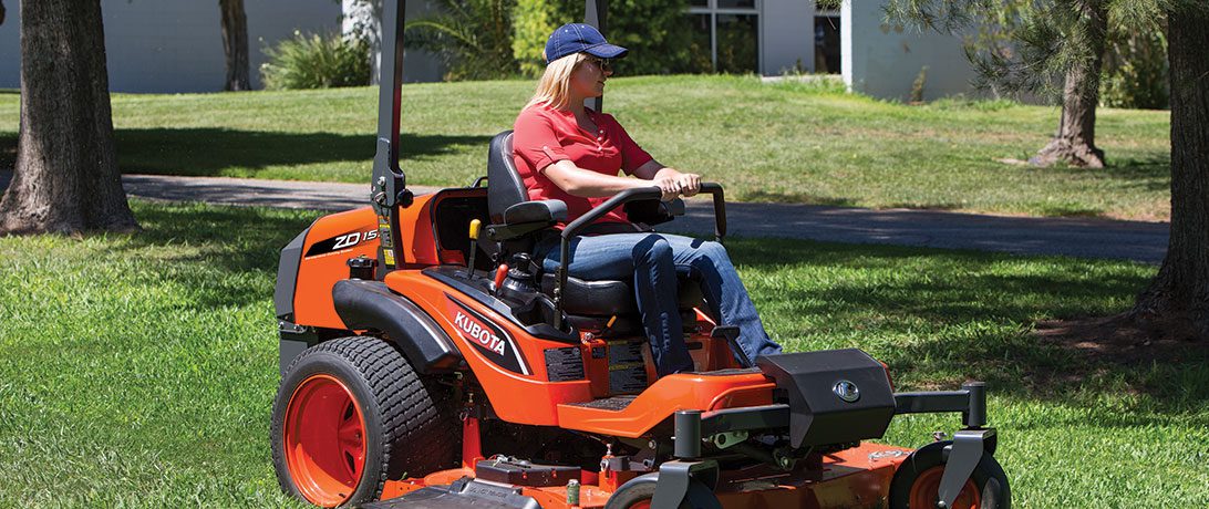 Person driving a Kubota zero-turn mower in the grass surrounding a building.