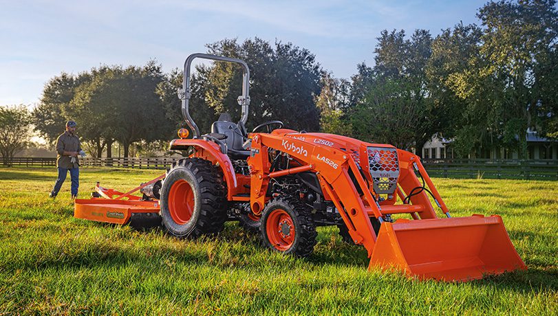 Person walking next to a Kubota L2502 Tractor in a field with trees in the background.